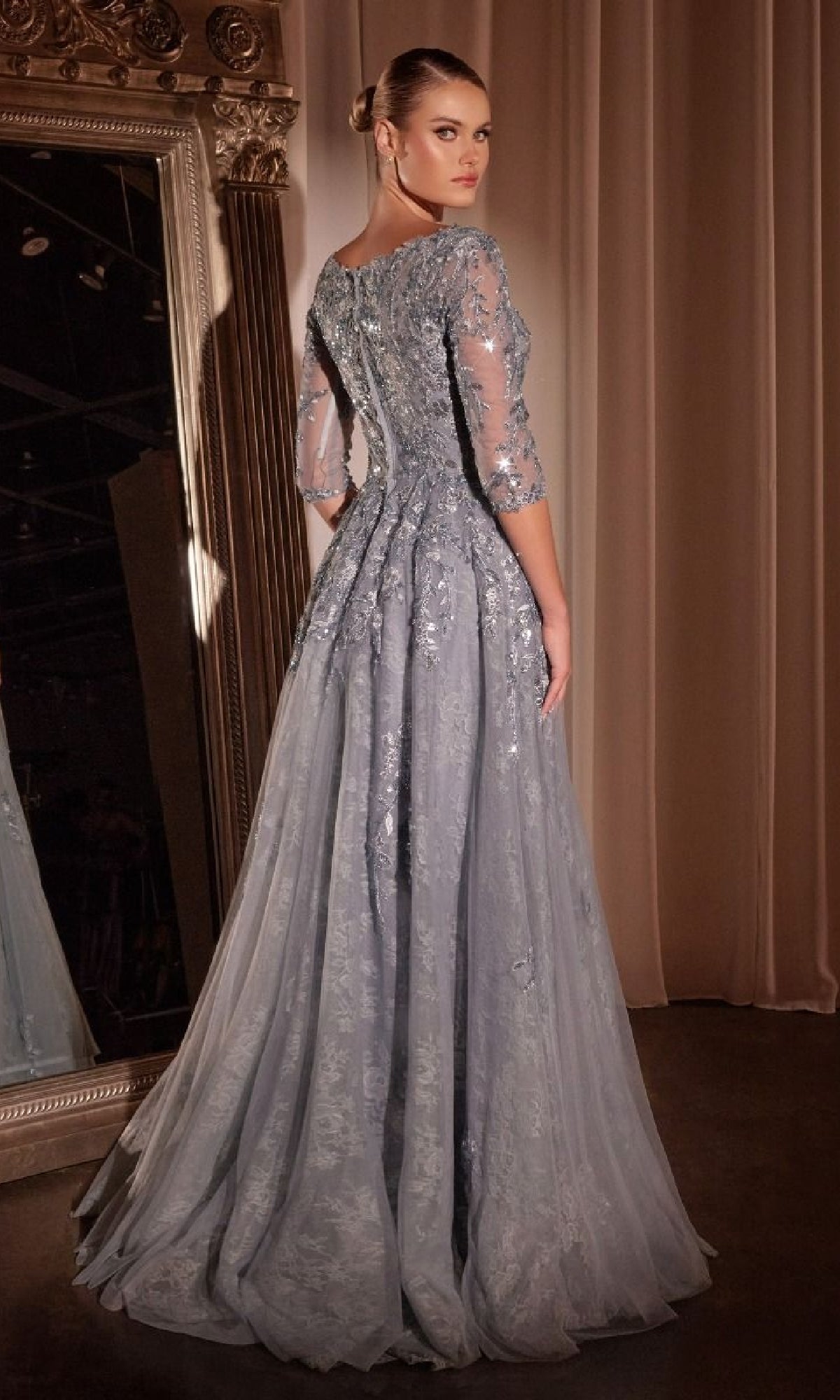 A woman in the Ladivine CK2401 long sleeve blue lace formal gown stands before a large ornate mirror, looking over her shoulder.